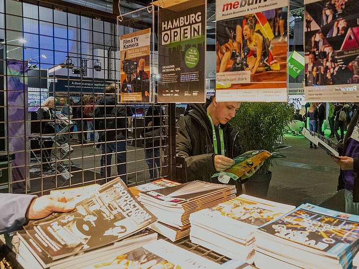 Blick auf den Fachpressestand der HAMBURG OPEN. Von links hält eine Person eine Zeitschrift ins Bild.