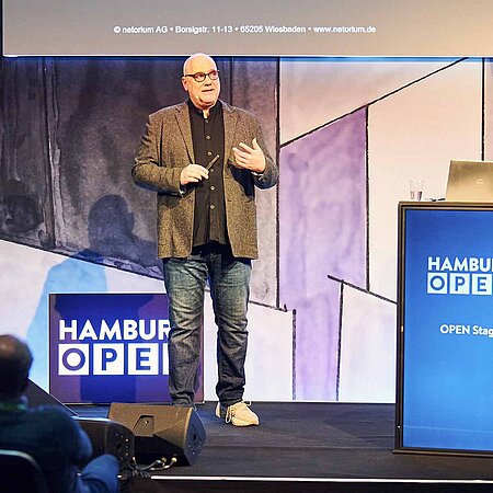 Speaker steht neben seinem Pult auf der OPEN Stage und präsentiert vor Publikum.