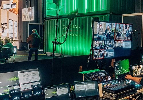 The recording area of ​​the forum can be seen in the foreground. Two men sit in front of monitors and observe what is happening on stage and the quality of the recording. The stage can be seen in the background.