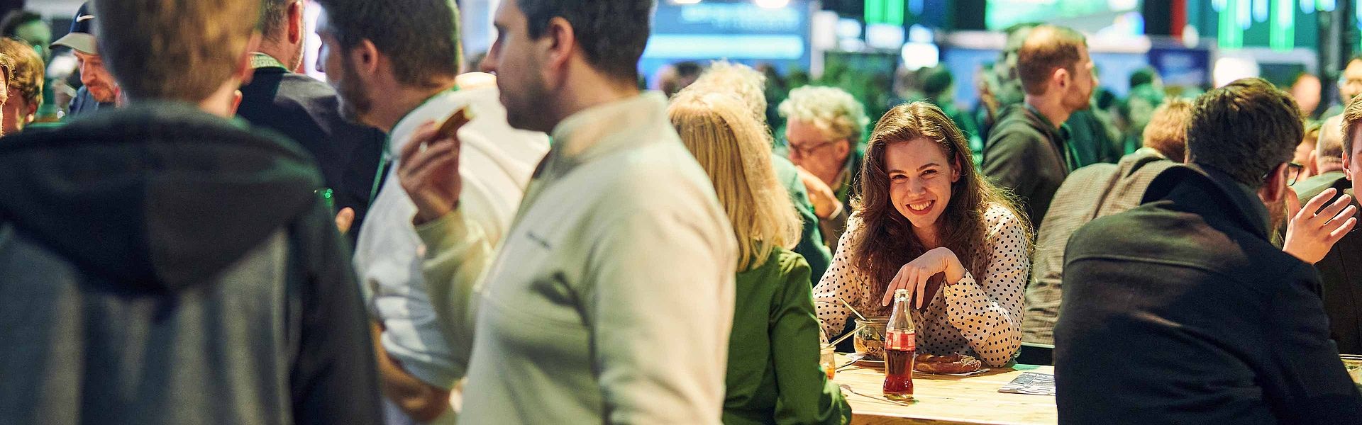 Blick in die Menschengruppe auf der Get-Together. Eine junge Frauch lächelt in die Kamera. Neben ihr befinden sich verschiedene Gruppen im Gespräch. Die Leute trinken und essen dabei.