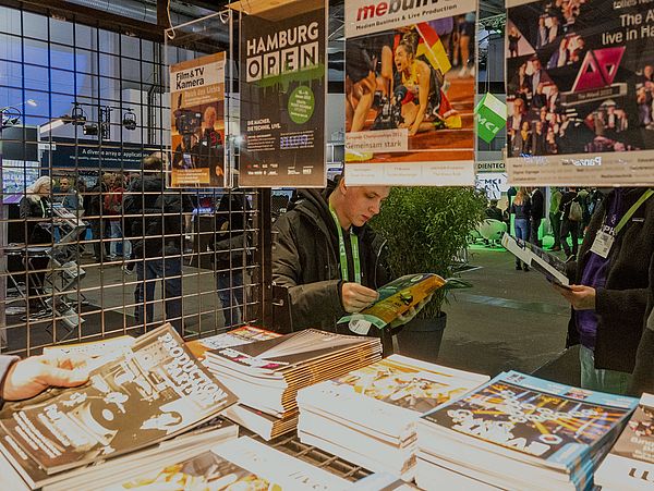 View of the trade press stand at the HAMBURG OPEN. From the left a person holds a magazine into the picture.