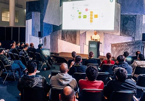   View over the heads of the audience onto the stage. A speaker gives a lecture. There is a graph shown on the graphic above him.