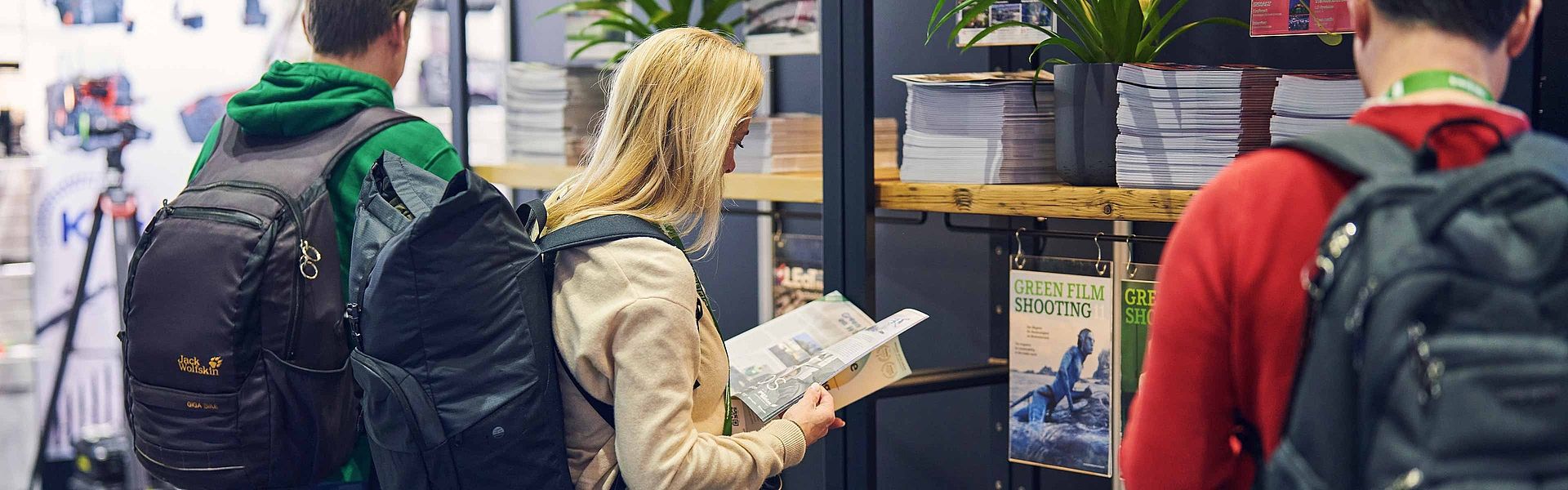 Eine Frau liest auf der Messe in einer Zeitschrift. Zwei Männer stehen links und rechts neben ihr und stöbern im Zeitschriftenregal.